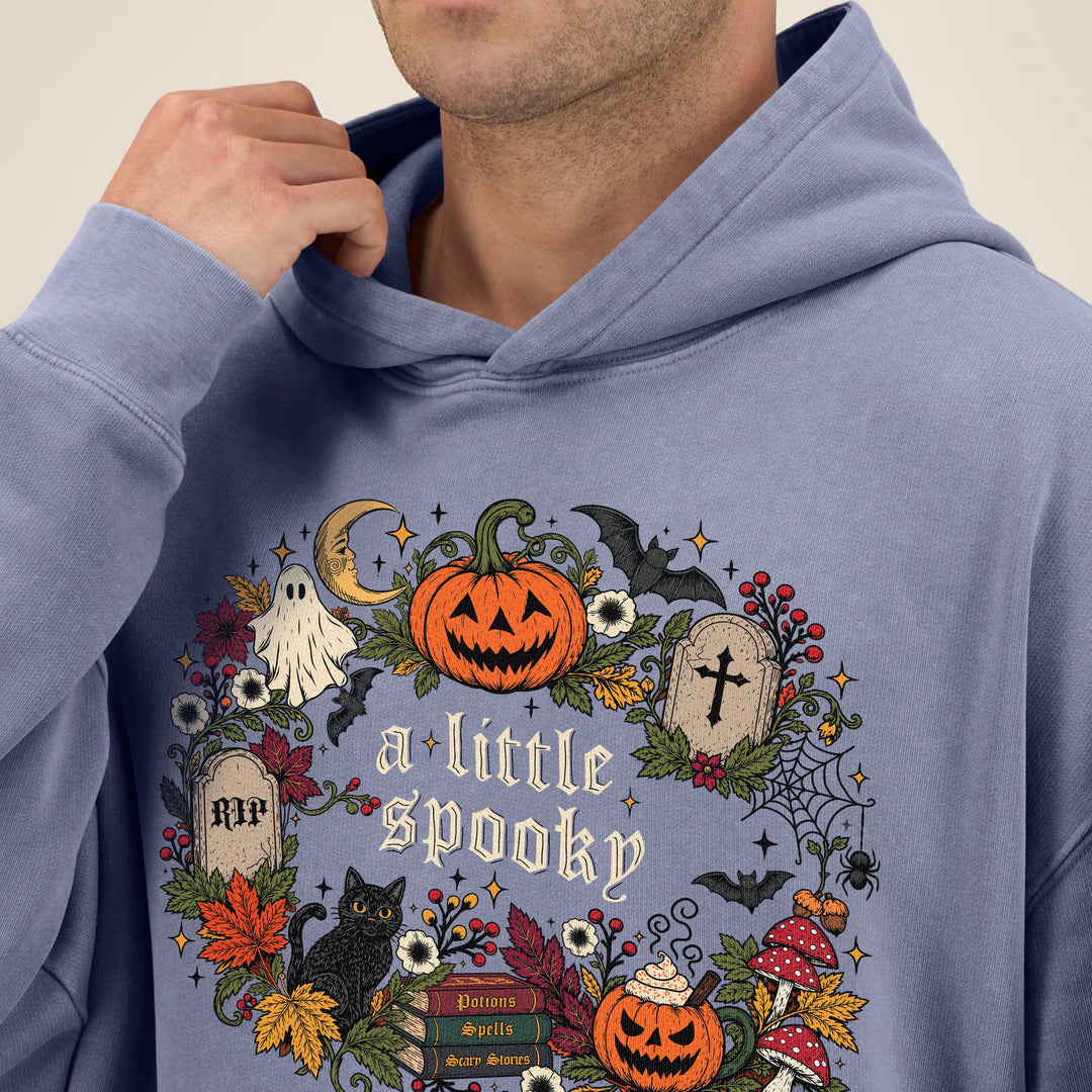 A man wearing a dusty blue hoodie with a detailed Halloween graphic on the chest that reads 'a little spooky' and features pumpkins, ghosts, bats, and autumn leaves.
