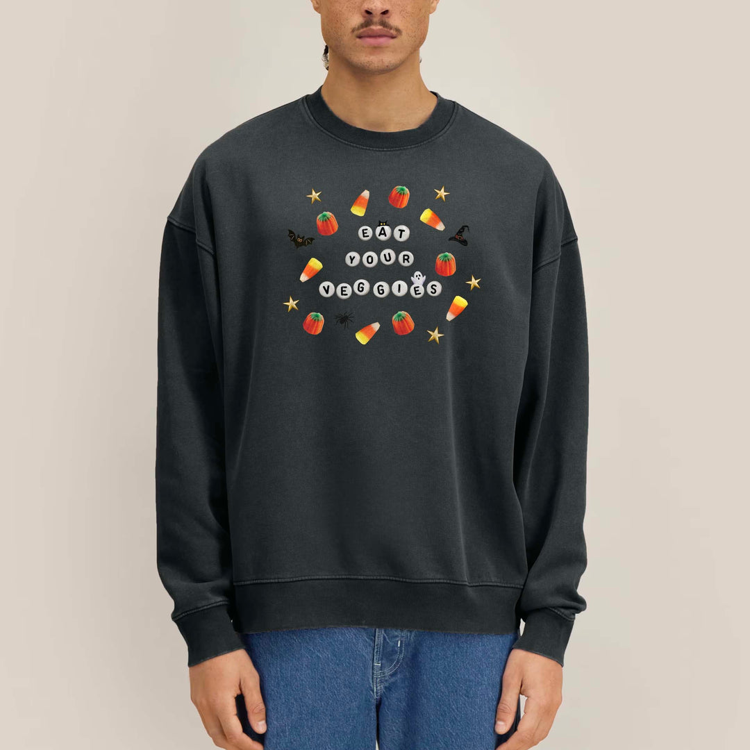 A man wearing a dark charcoal gray crewneck sweatshirt with a colorful graphic on the chest that reads 'Candy Corn Celebration' surrounded by illustrations of candy corn, lollipops, and stars. He is wearing blue jeans against a solid light beige background.