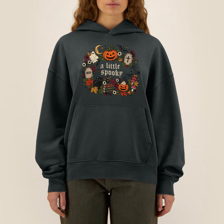 A woman wearing a dark charcoal gray hoodie with a colorful Halloween graphic on the chest that reads 'a little spooky' surrounded by pumpkins, ghosts, and autumn leaves, set against a plain beige background.
