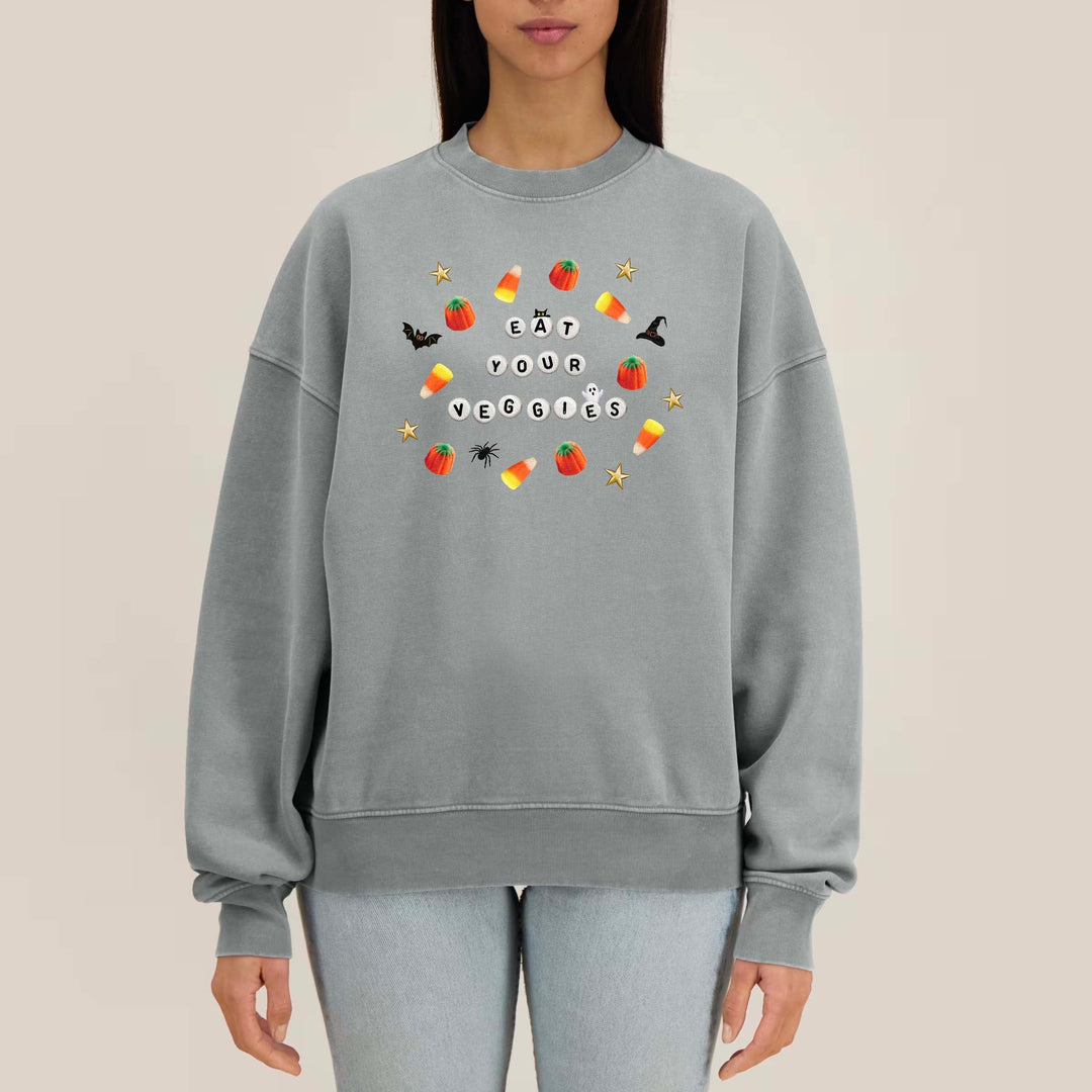 A woman modeling a light gray crewneck sweatshirt with a colorful Halloween-themed graphic that reads 'eat your demons' surrounded by candy corn, ghosts, and bats, against a solid beige background.