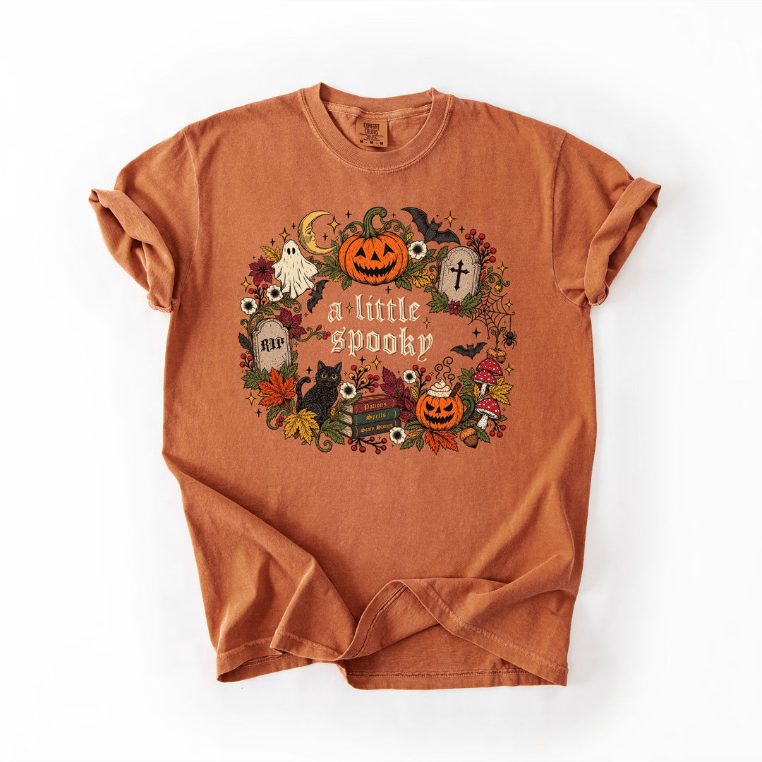 An orange crewneck t-shirt with a distressed texture, featuring a circular Halloween graphic on the front with pumpkins, ghosts, a cat, and the text 'a little spooky'.