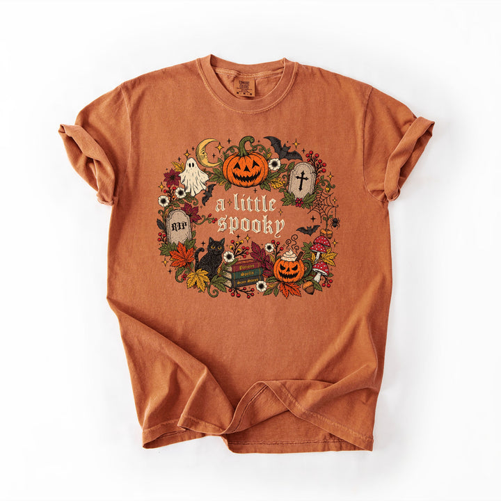 An orange crewneck t-shirt with a distressed texture, featuring a circular Halloween graphic on the front with pumpkins, ghosts, a cat, and the text 'a little spooky'.