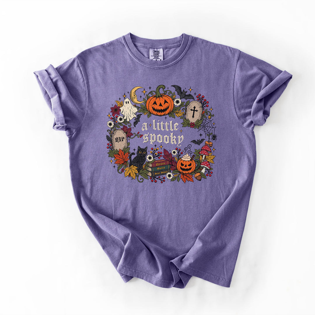 A lavender purple short-sleeve t-shirt with a colorful Halloween graphic on the front that reads 'a little spooky'. The graphic features jack-o'-lanterns, a ghost, a cat, tombstones, and a witch hat.