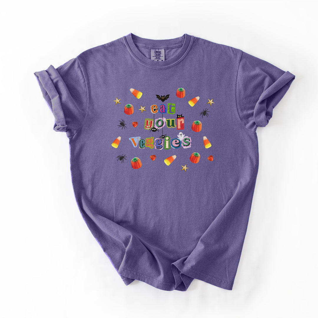 A purple short-sleeve t-shirt with a colorful graphic on the chest that reads 'eat your vegetables' surrounded by Halloween-themed illustrations like candy corn, pumpkins, spiders, and bats, laid flat on a white background.