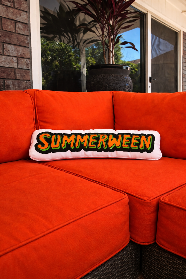 Orange outdoor sofa with a 'Summerween' pillow against a brick wall and glass door background.