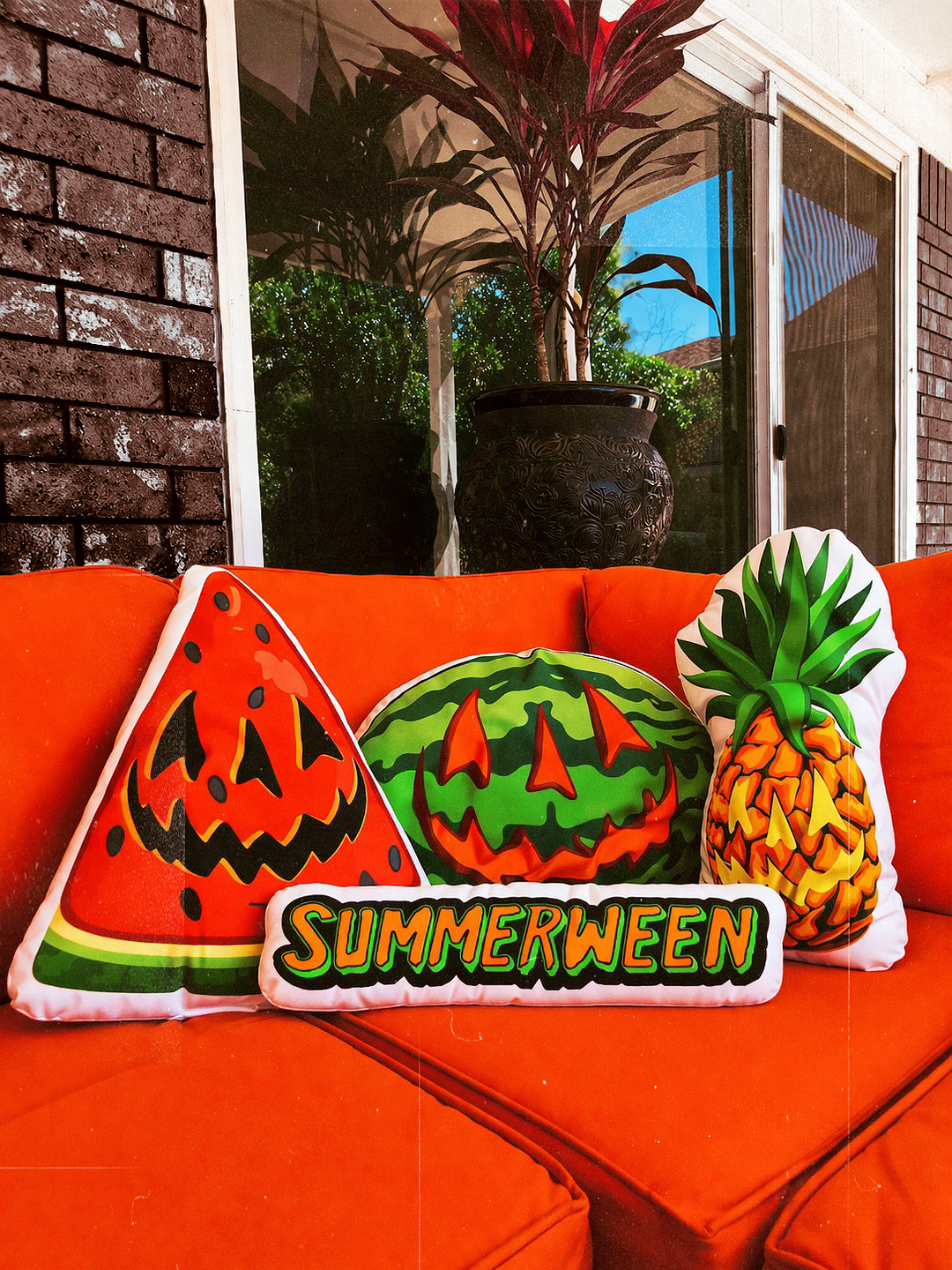 Decorative pillows shaped like watermelon, pineapple, and jack-o'-lantern on an orange couch with 'Summerween' text.